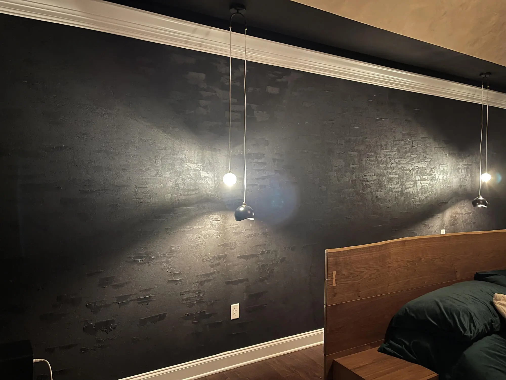 Modern bedroom featuring a dramatic black textured accent wall with crown molding and hanging pendant lights above a wooden platform bed with dark bedding