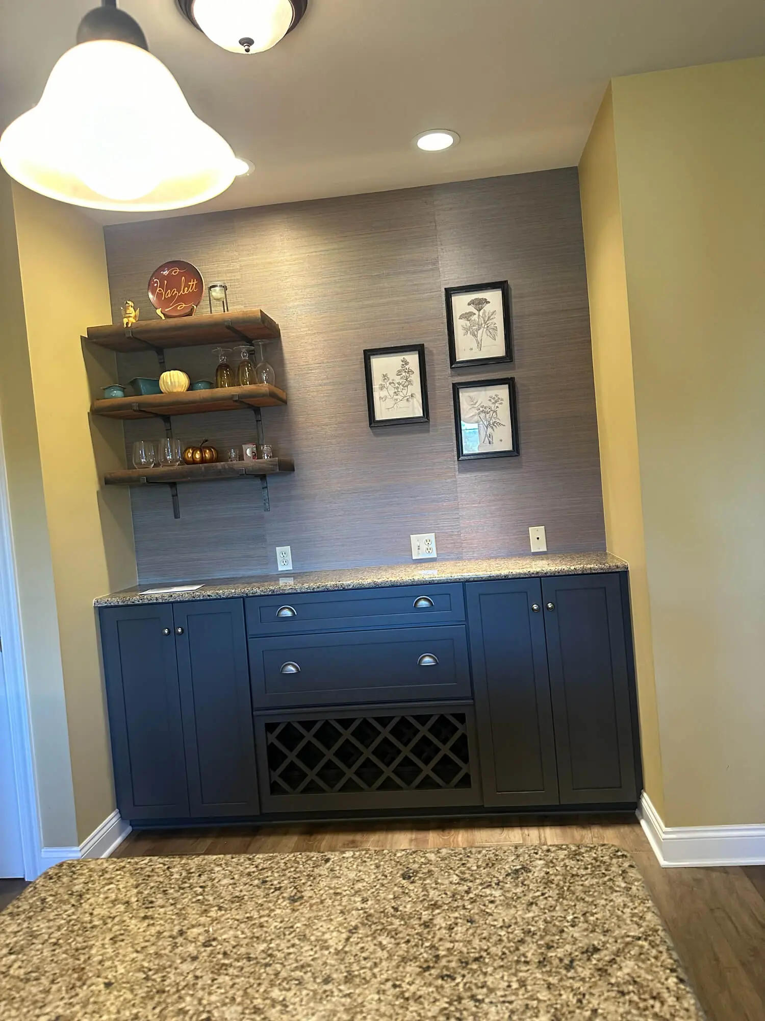 Dark navy built-in bar cabinet with granite countertop, wine rack, and floating wood shelves styled with décor accents against a textured accent wall