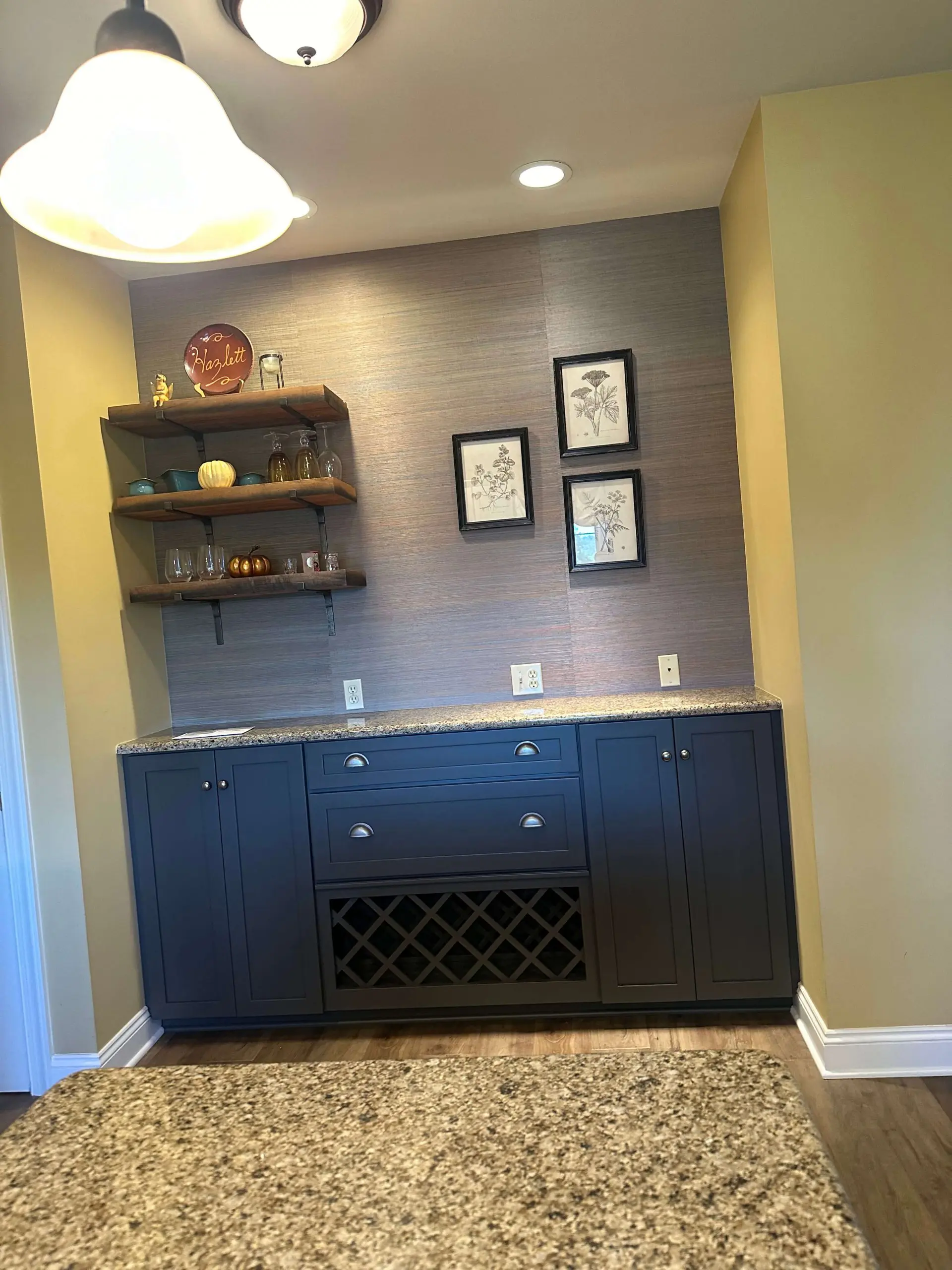 Modern home bar featuring dark navy cabinetry, granite countertops, and Martzalls custom surface. organized rustic wood shelves against a textured grey grasscloth wallpaper.
