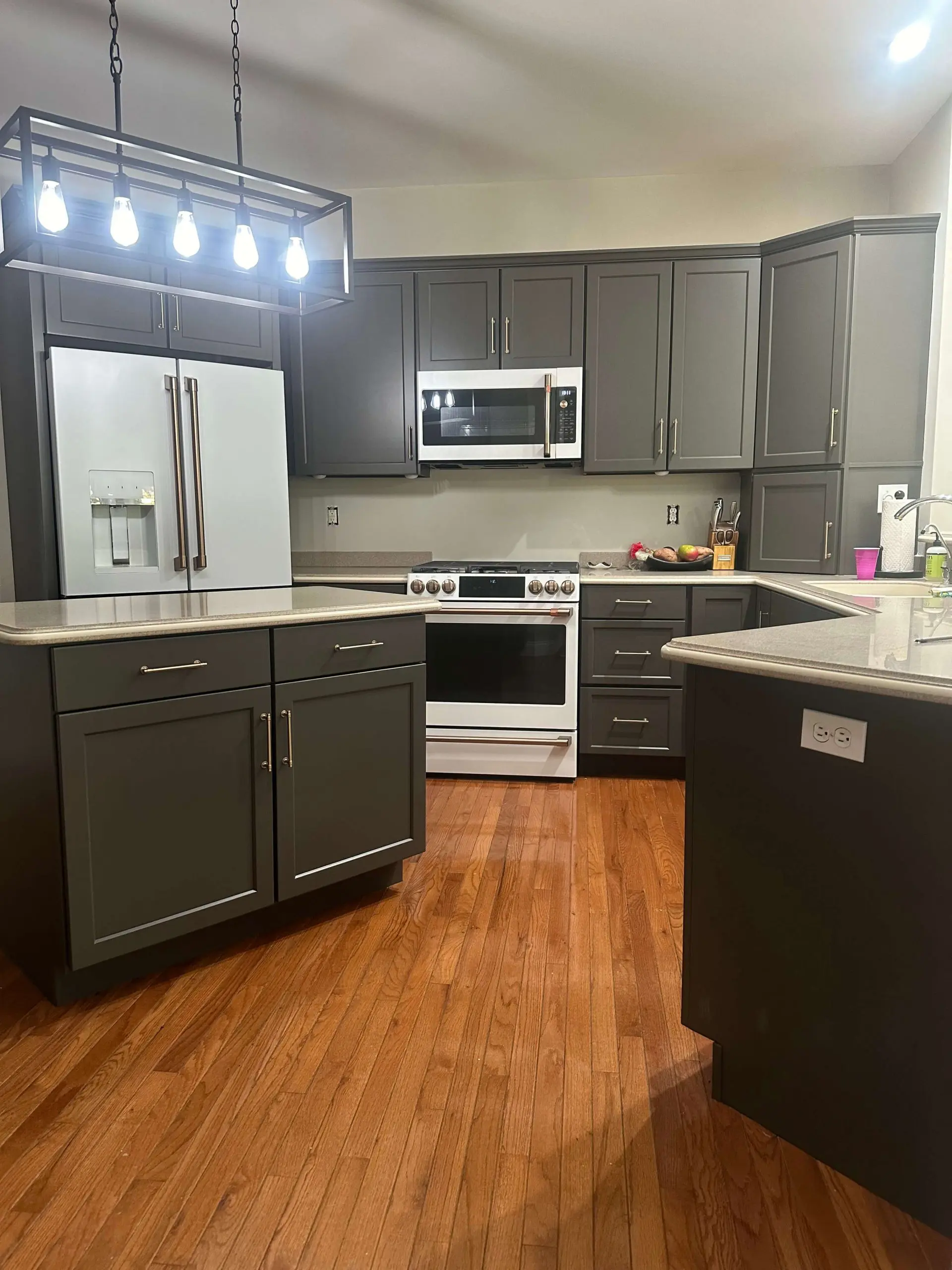 Martzall's Custom Surfaces showcases a bold kitchen design with charcoal grey shaker cabinets, light quartz countertops, and modern white appliances illuminated by a black industrial frame chandelier.