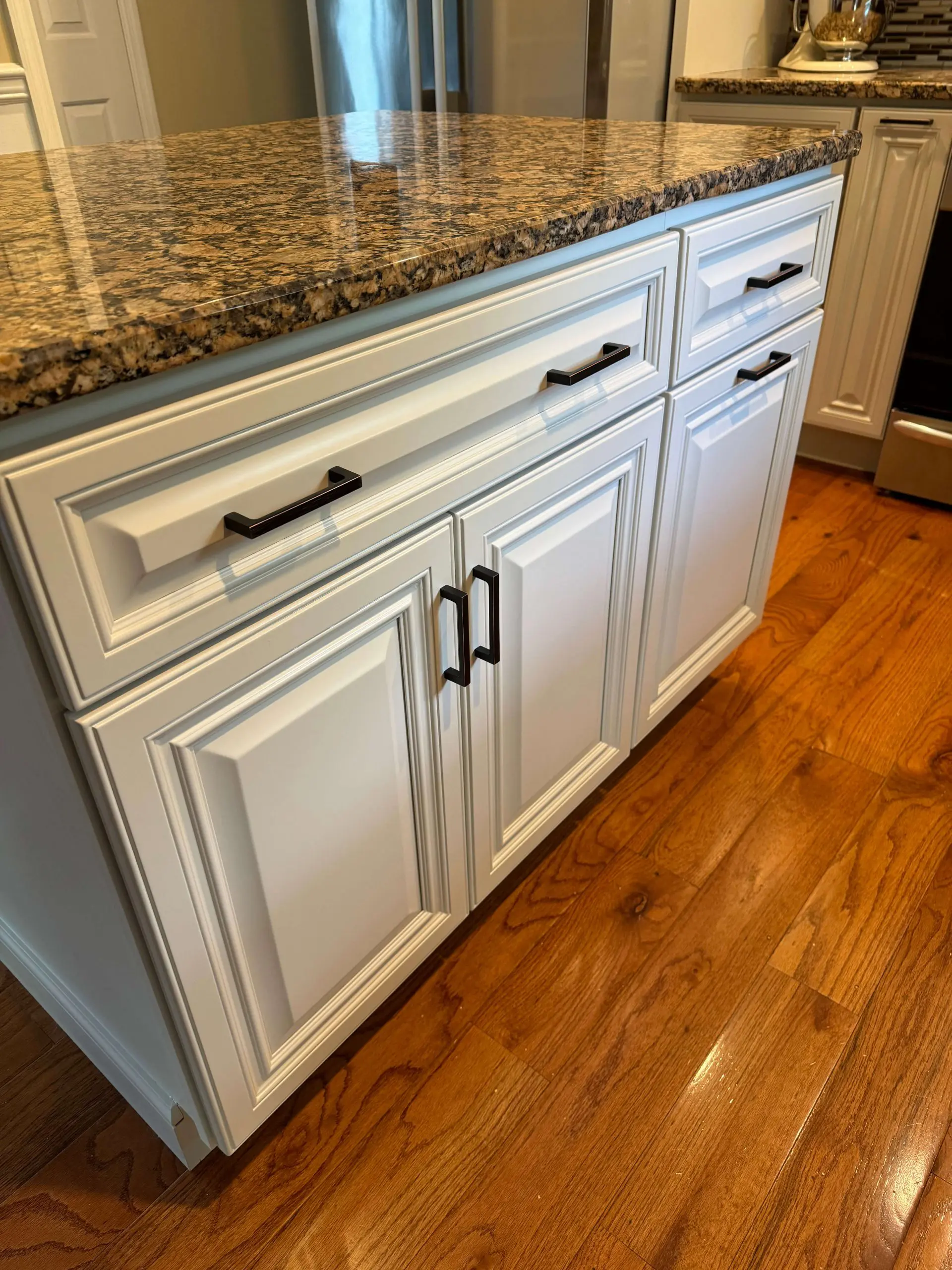 Custom kitchen island by Martzall's Custom Surfaces featuring elegant off-white raised panel cabinetry, matte black hardware, and polished granite countertops on hardwood flooring.