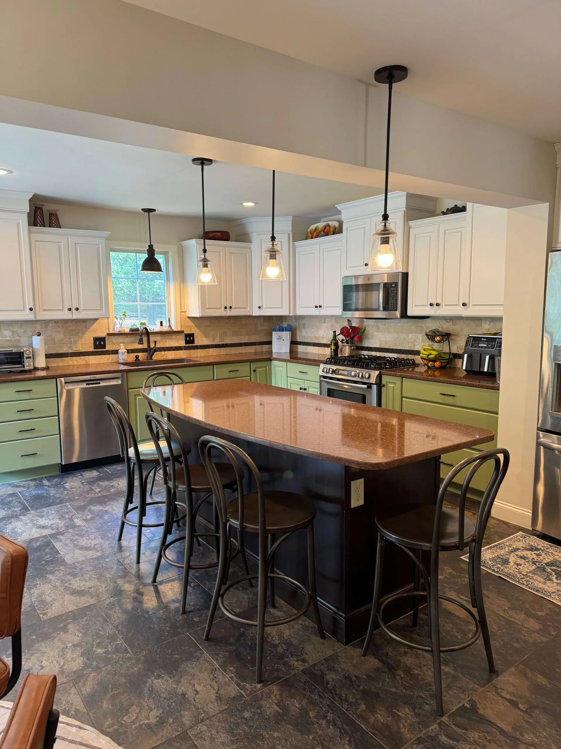 Spacious kitchen remodel from Martzall's Custom Surfaces showcasing sage green base cabinets, polished granite island, and contemporary industrial pendant lighting.