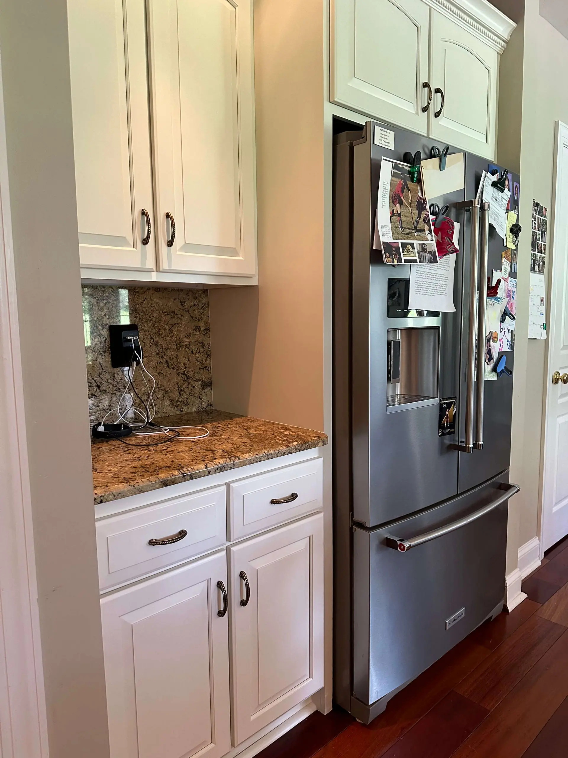Stainless steel French door refrigerator alongside white kitchen cabinets and a granite-topped charging station nook.