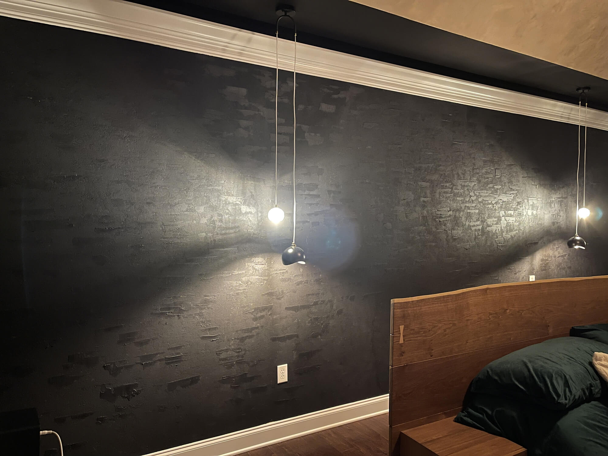 Modern bedroom featuring a dramatic black textured accent wall with crown molding and hanging pendant lights above a wooden platform bed with dark bedding